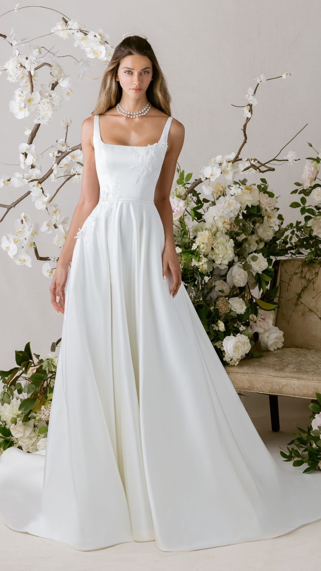 Elegantes Brautkleid von Morilee mit schlichten Trägern, verziert mit Blumenarrangements im Hintergrund.