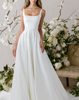 Elegantes Brautkleid von Morilee mit schlichten Trägern, verziert mit Blumenarrangements im Hintergrund.