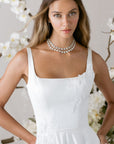 Elegantes Brautkleid von Morilee mit schlichten Trägern, verziert mit Blumenarrangements im Hintergrund.