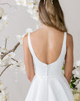 Elegantes Brautkleid von Morilee mit schlichten Trägern, verziert mit Blumenarrangements im Hintergrund.