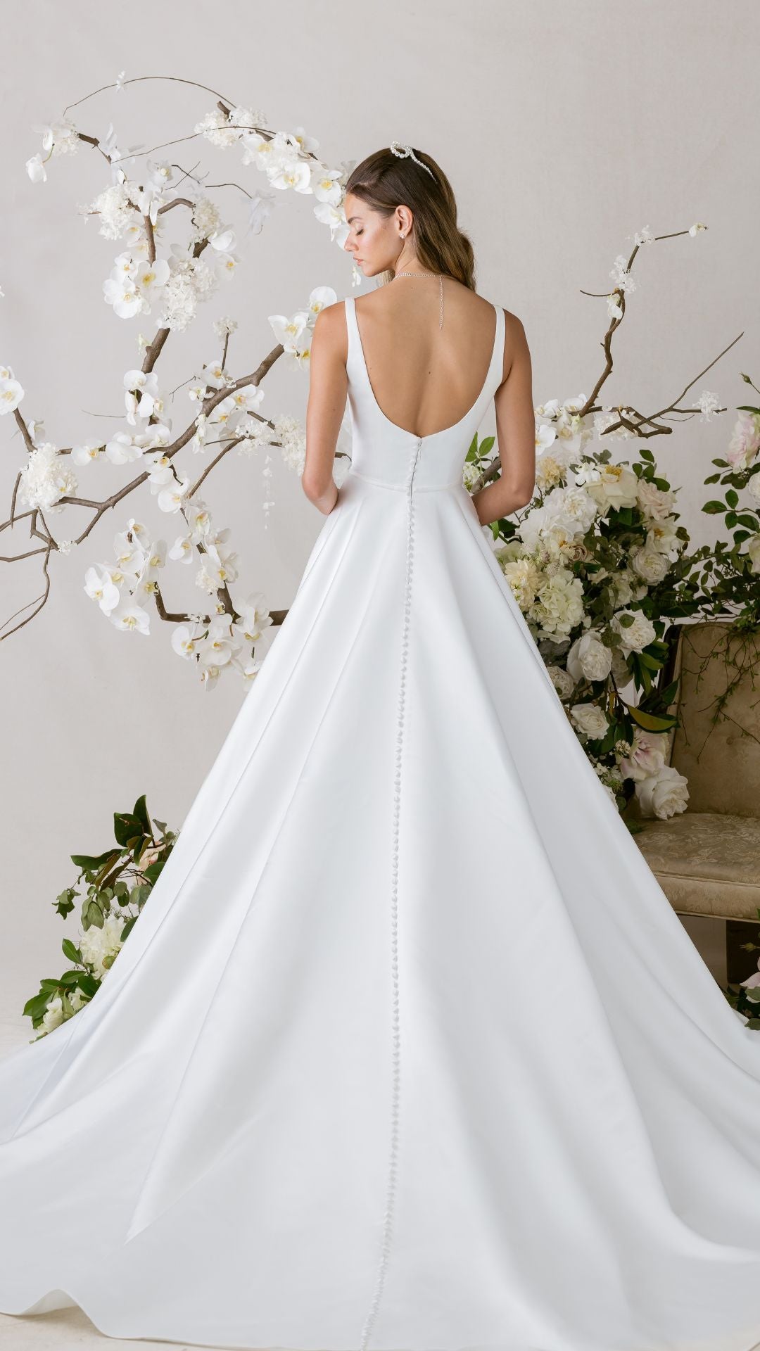 Elegantes Brautkleid von Morilee mit schlichten Trägern, verziert mit Blumenarrangements im Hintergrund.