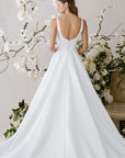Elegantes Brautkleid von Morilee mit schlichten Trägern, verziert mit Blumenarrangements im Hintergrund.